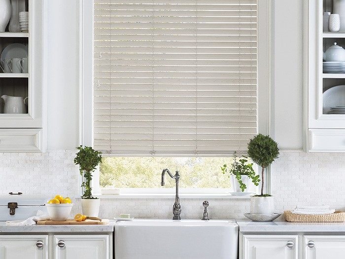 Kitchen with beautiful blinds above a French farmhouse porcelain sink with white cabinetry, marble kitchen counter tops and white tiled black splash.